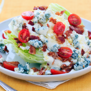 Wedge of iceberg lettuce with blue cheese dressing, sliced cherry tomatoes, bacon crumbles, and blue cheese on a white salad plate