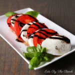 White plate with sliced tomato, mozzarella cheese with a drizzle of balsamic glaze and a garnish of fresh basil leaves.