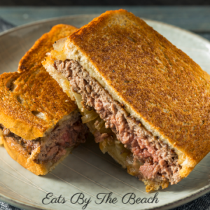 A classic patty melt of grilled sourdough bread, ground beef patty, caramelized onion, and cheese on a white plate