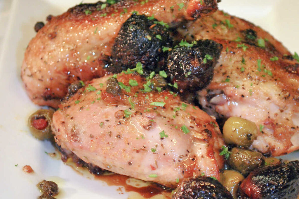 Closeup view of three pieces of bone-in Chicken Marbella, with pan juices, prunes, and green olives, and garnished with a sprinkling of chopped fresh parsley.