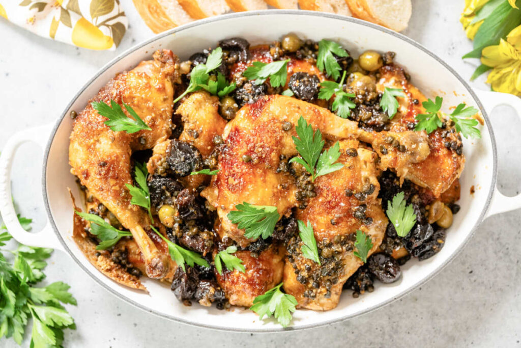 Chicken Marbella in a white casserole dish. Cut up bone-in chicken stewed with white wine, prunes, capers, garlic, oregano, and spices. Garnished with whole parsley leaves.