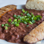 White bowl of creamy and spicy red beans and rice served with a side of grilled Andouille sausage and French bread, garnished with sliced scallions