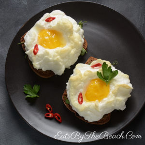 Trendy brunch dish. Plate of two fluffy, parmesan egg white clouds cradling a sunny, soft-baked yolk. Served on a slice of toasted brown bread and garnished with sliced red chiles and parsley.