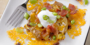 A plate with a baby potato that has been boiled, smashed into a patty and then browned in garlic butter. It's then baked with cheddar cheese and bacon, then garnished with sour cream and chopped green onion. This loaded smashed potato has all the flavors of your favorite loaded baked potato.