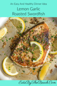 A skillet with Lemon Garlic Roasted Swordfish, garnished with lemon slices and chopped chives in a lemon, wine sauce.