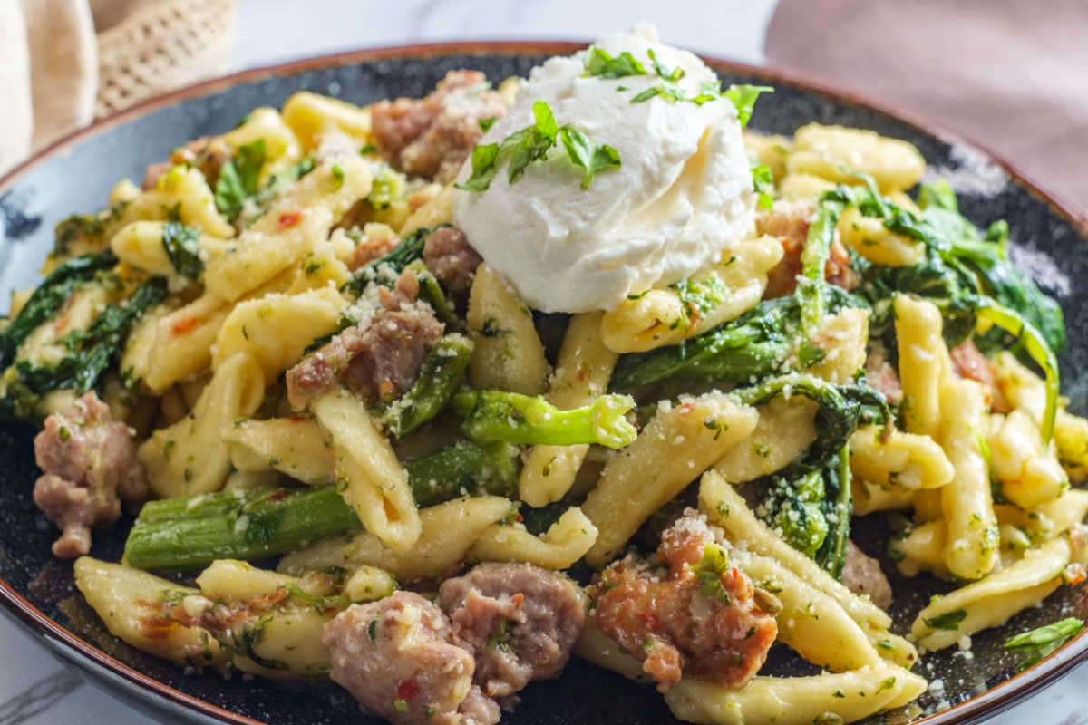 A brown bowl of homemade cavatelli with sweet Italian sausage and broccoli rade, garnishe with a dollop of whole milk ricotta chese.