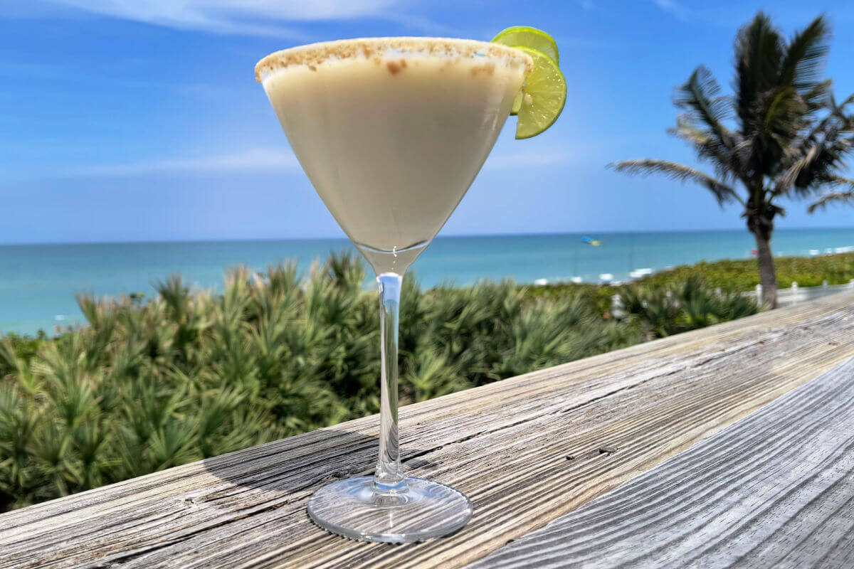 A sweet, creamy Key Lime Pie Martini on a wooden table by the beach.