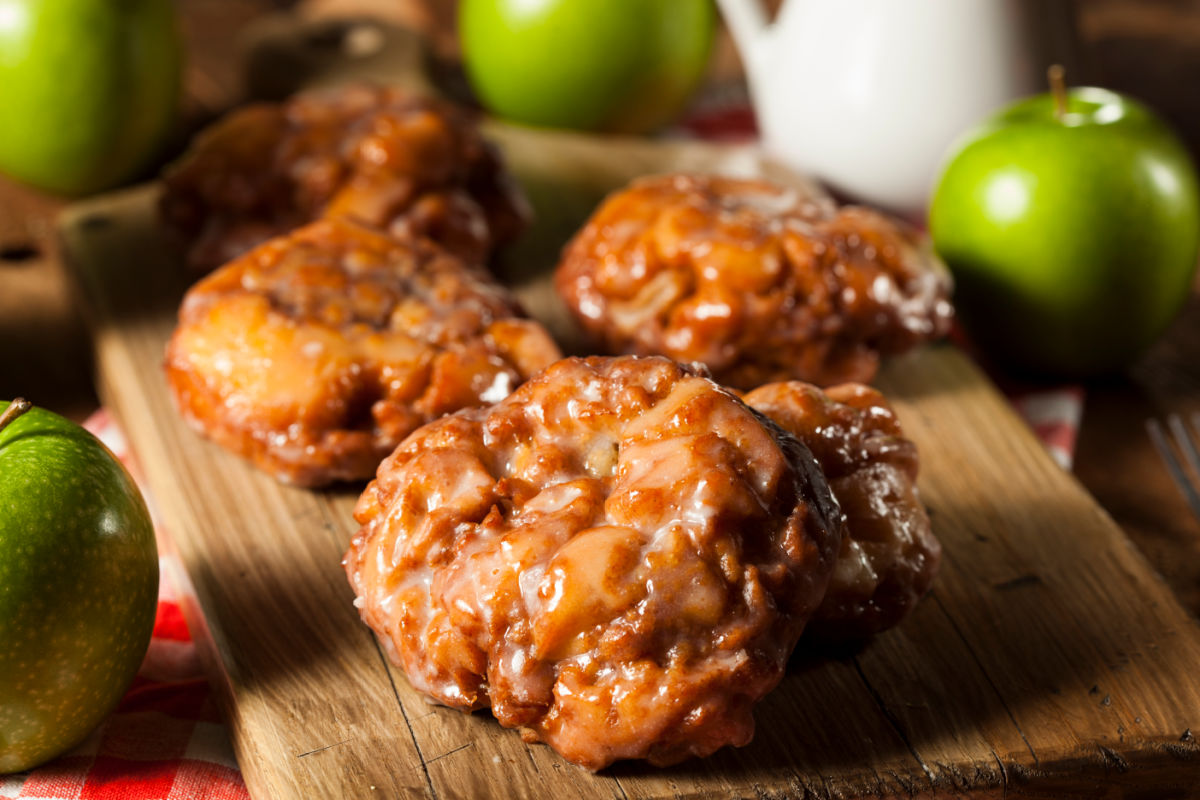 Wooden board with homemade glazed cinnamon apple fritters.  Beside the board are fresh Granny Smith apples and a glass of milk.