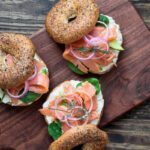 Wooden board with easy California Lox Bagel Sandwiches - Everything bagel with smoked salmon, spinach, red onions, avocado and cream cheese.