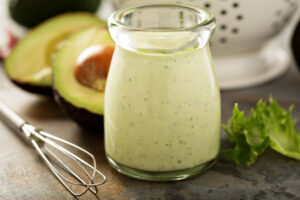 Jar of homemade avocado ranch dressing with a white colander of salad greens and avocado halves beside it.