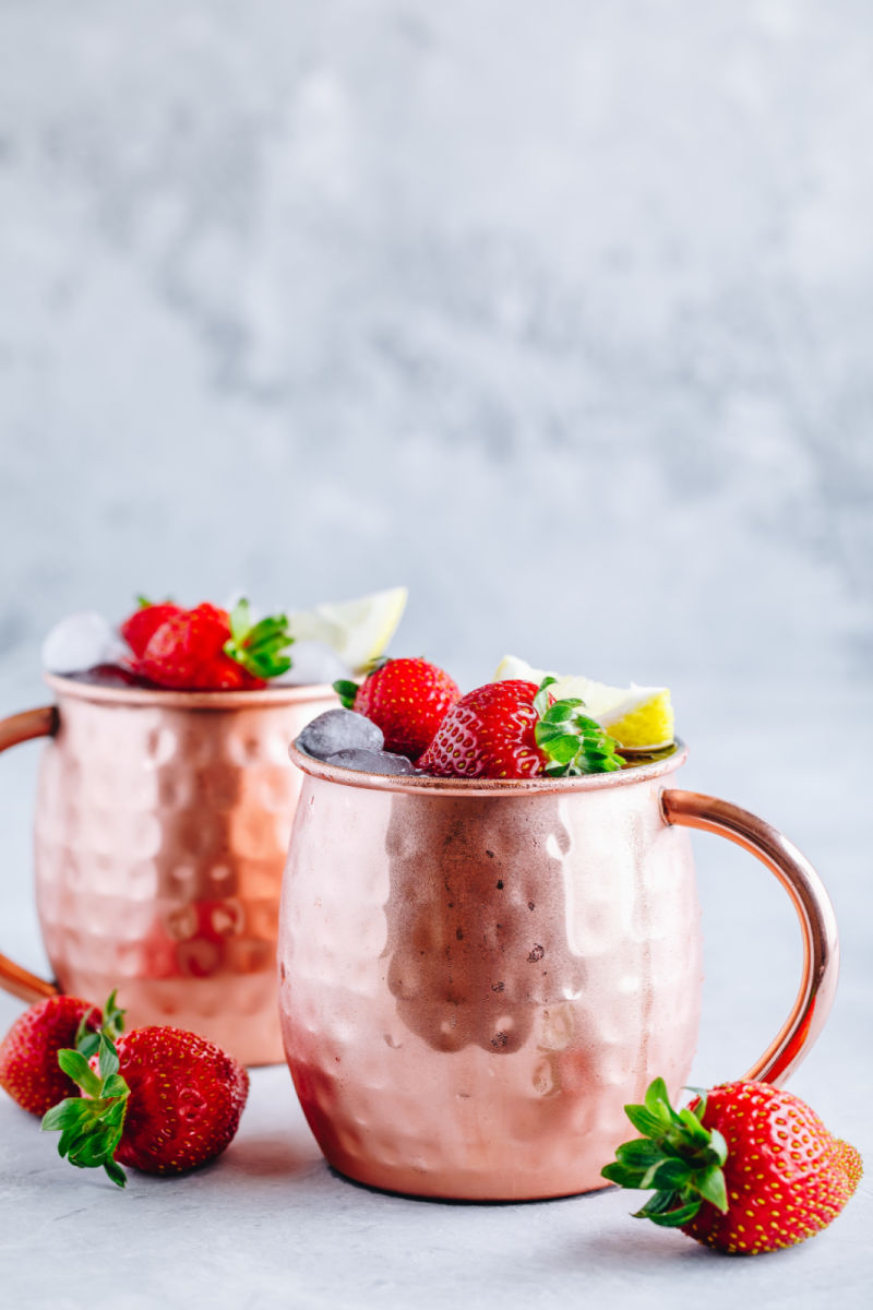 2 copper Moscow Mule mugs with strawberry Moscow Mule cocktails, garnished with whole strawberries and a lemon wedge.