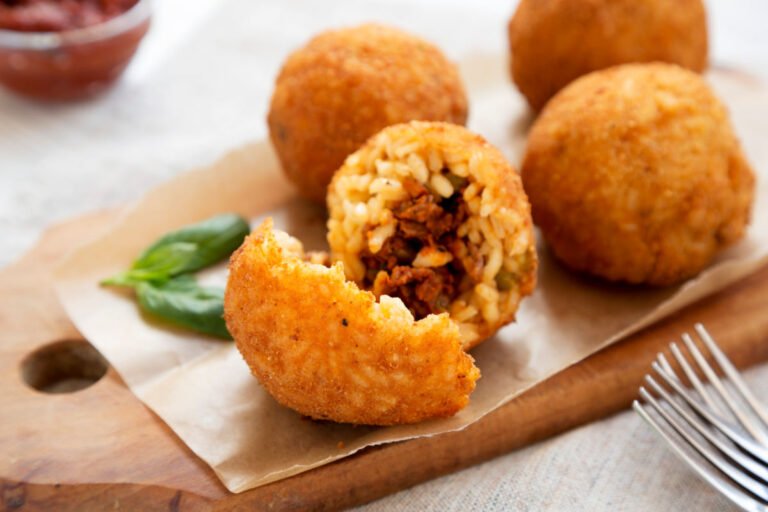 Wooden serving board with golden, deep-fried balls of Arancini di Riso and one ball showing the inside with ground beef and peas..