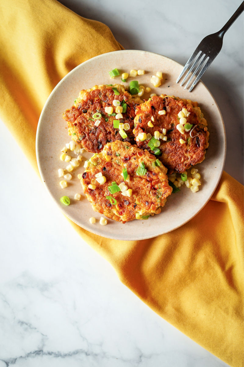 White plate with 3 sweet corn fritters and garnished with fresh corn kernels and sliced scallion, sitting on a yellow napkin on a white marble slab.