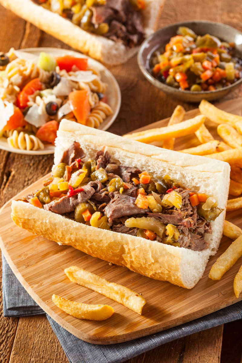 Top view of Authentic Italian Beef Sandwiches served on wooden boards with a side of french fries and a bowl of hot Giardiniera.