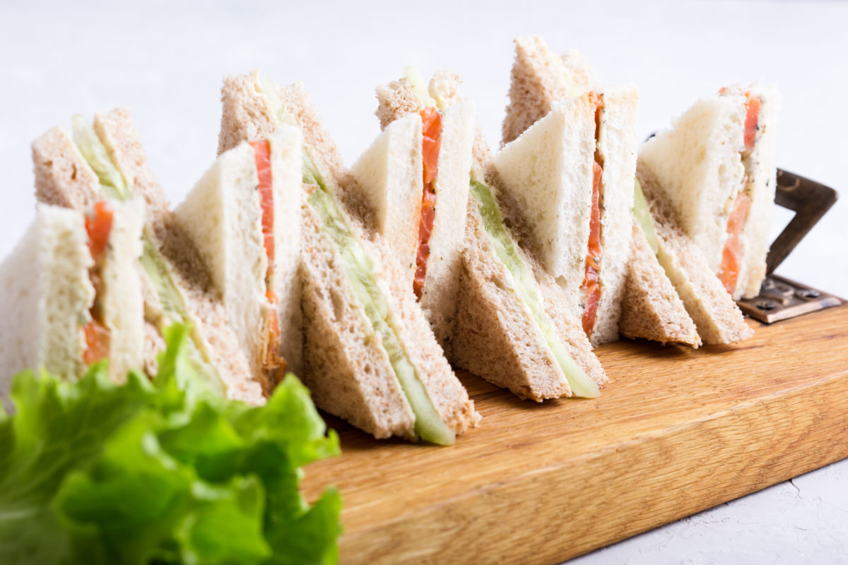 Wooden platter of The Queen's Favorite Smoked Salmon And Cucumber Tea Sandwiches cut into triangles and stacked decoratively.