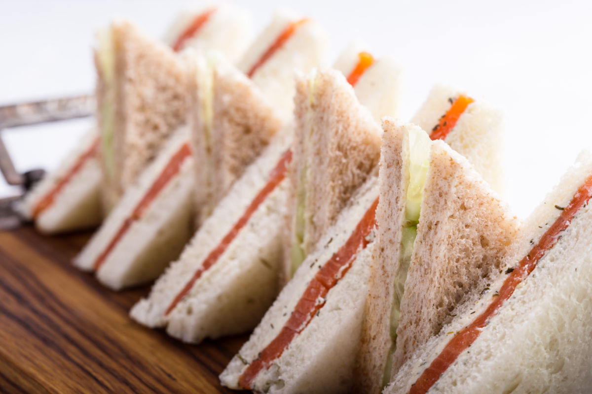 Wooden platter of The Queen's Favorite Smoked Salmon And Cucumber Tea Sandwiches cut into triangles and stacked decoratively.