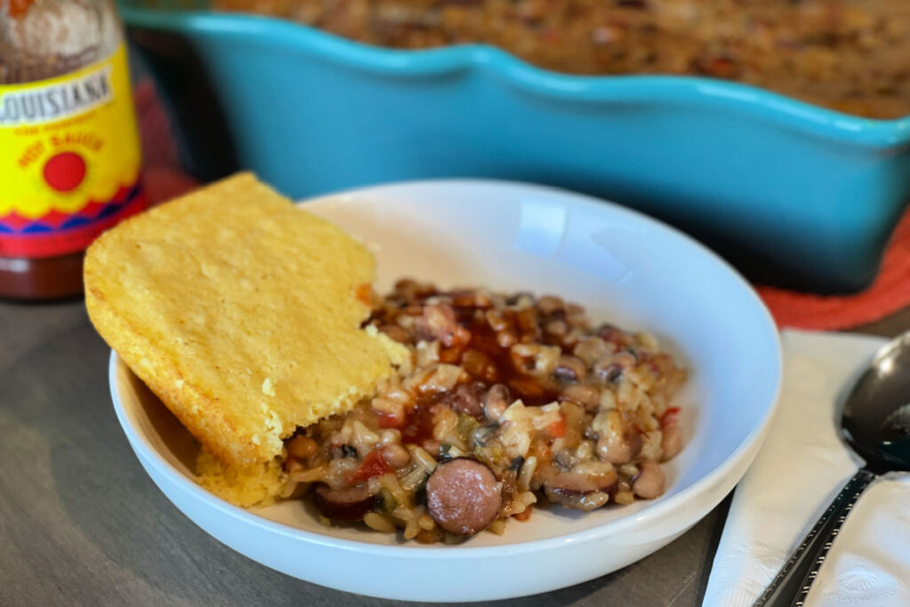 A white bowl with a serving of New Year's Hoppin John Casserole-black eyed peas, andouille sausage, dirty rice and aromatic veggies baked in a creamy sauce. It's served with a side of cornbread.