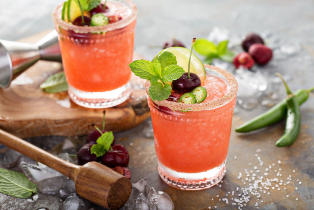 Two short cocktail glasses with spicy Cherry Chili Lime Margaritas garnished with jalapeno slices, fresh cherry halves, lime slices, and a mint sprig.