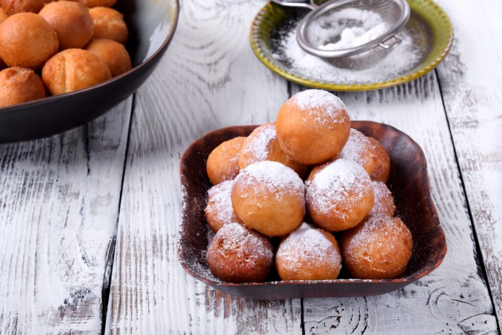 Wide view of a bowl f stacked old-fashioned beignets beside a bowl of powdered sugar and a fine mesh sieve.