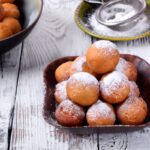 Wide view of a bowl f stacked old-fashioned beignets beside a bowl of powdered sugar and a fine mesh sieve.