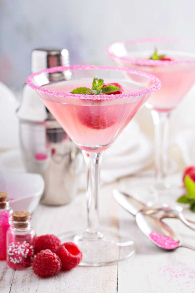 Two pink copycat Pump-Tini martinis on a white lined table with raspberries, a cocktail shaker, sanding sugar, and a spoon as garnish.