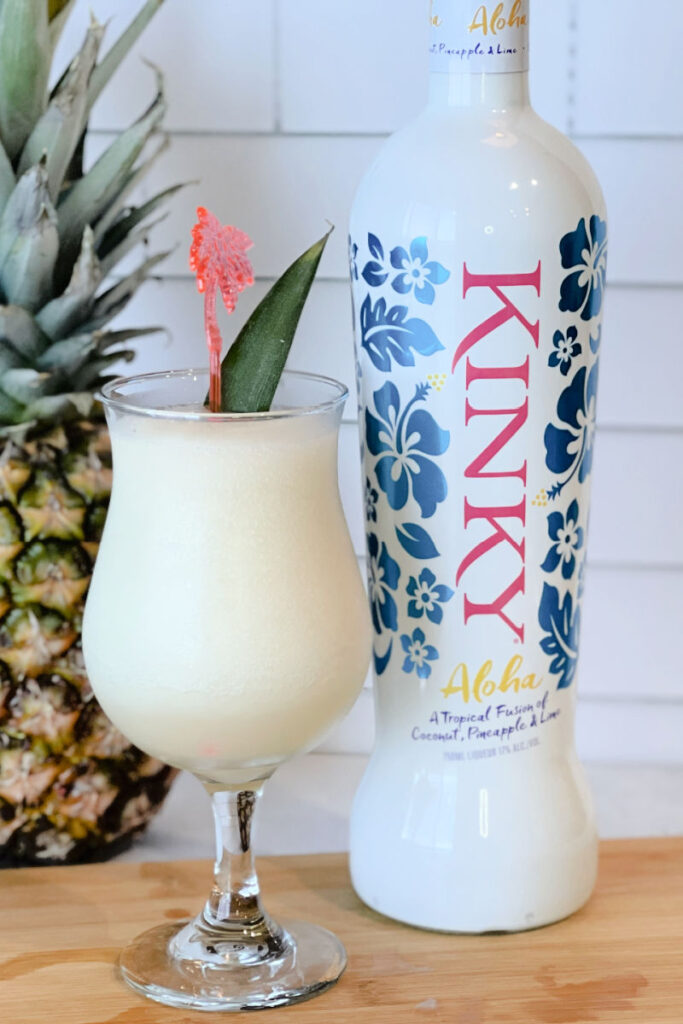 Side view of a Hurricane glass with a Kinky Colada cocktail sitting beside a bottle of Kinky Aloha liqueur and a fresh pineapple.