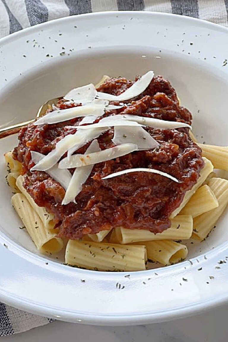 White bowl of Braised beef ragu served over ziti and garnished with Parmesan cheese shavings. Close up top view.