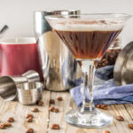 Martini glass with a Creamy tequila Espresso Martini on a blonde wooden board with a cocktail shaker and fresh coffee beans all around the glass.