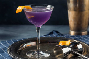 A Purple Rain Cocktail in a coupe glass with a twist of lemon on the rim on a tarnished silver tray with a shot glass and cocktail shaker in the background. Side view.