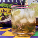 Closeup view of a beautiful cocktail glass of a Louisiana Libre cocktail made with King Cake soda, white rum, and lime juice. A lime slice garnish, Mardi Gras placemat, and a six-pack of Abita King Cake Soda completes the festive photo.