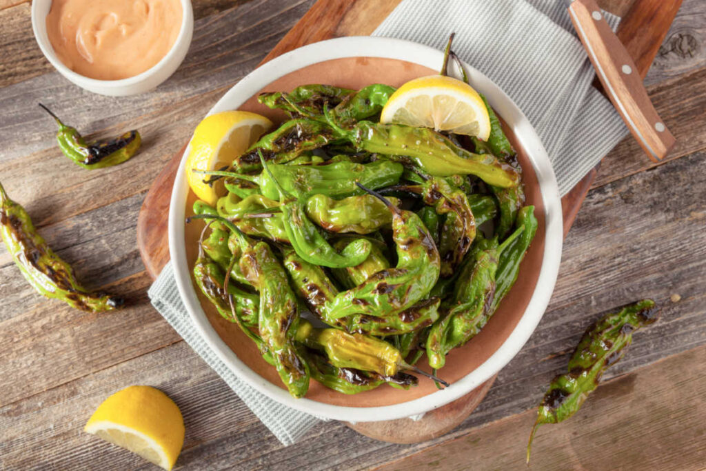 Rustic bowl with easy Cajun grilled shishito peppers garnished with lemon wedges and a side of Cajun Aioli sauce.