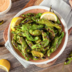 Rustic bowl with easy Cajun grilled shishito peppers garnished with lemon wedges and a side of Cajun Aioli sauce.