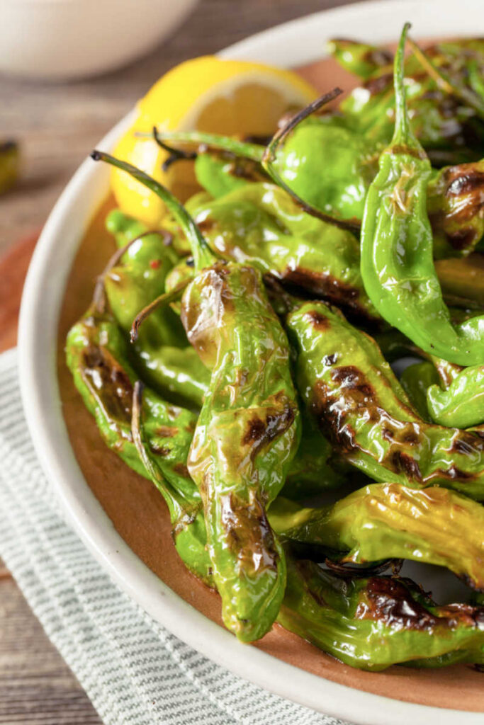 Rustic bowl with easy Cajun grilled shishito peppers garnished with lemon wedges and a side of Cajun Aioli sauce.