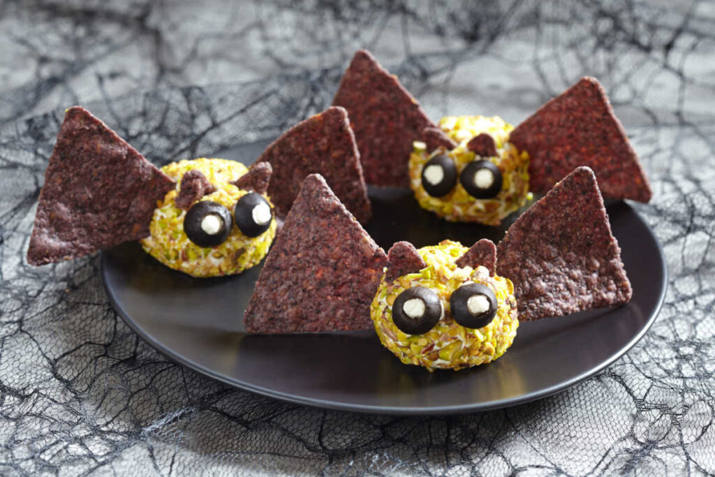 Black plate with 3 Batty Cheese Bites - a creamy cheese ball , rolled in chopped mpistachios and decorated to look like a bat with blue tortilla chi wings and ears. Black olives and goat cheese make the bat's eyes. Side view.