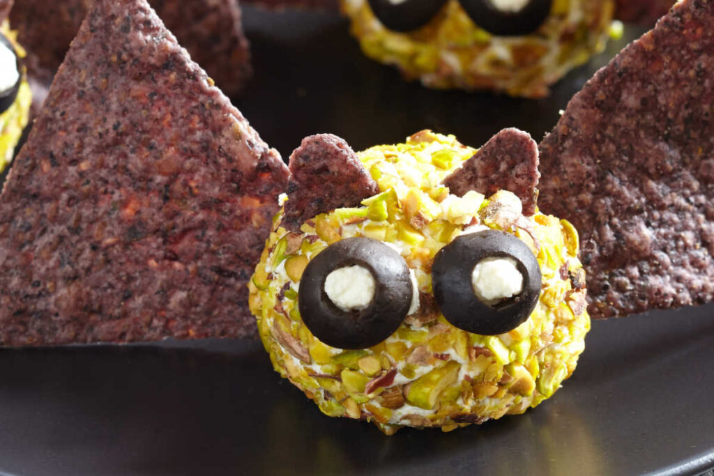 Black plate with 3 Batty Cheese Bites - a creamy cheese ball , rolled in chopped mpistachios and decorated to look like a bat with blue tortilla chi wings and ears. Black olives and goat cheese make the bat's eyes. Side view.