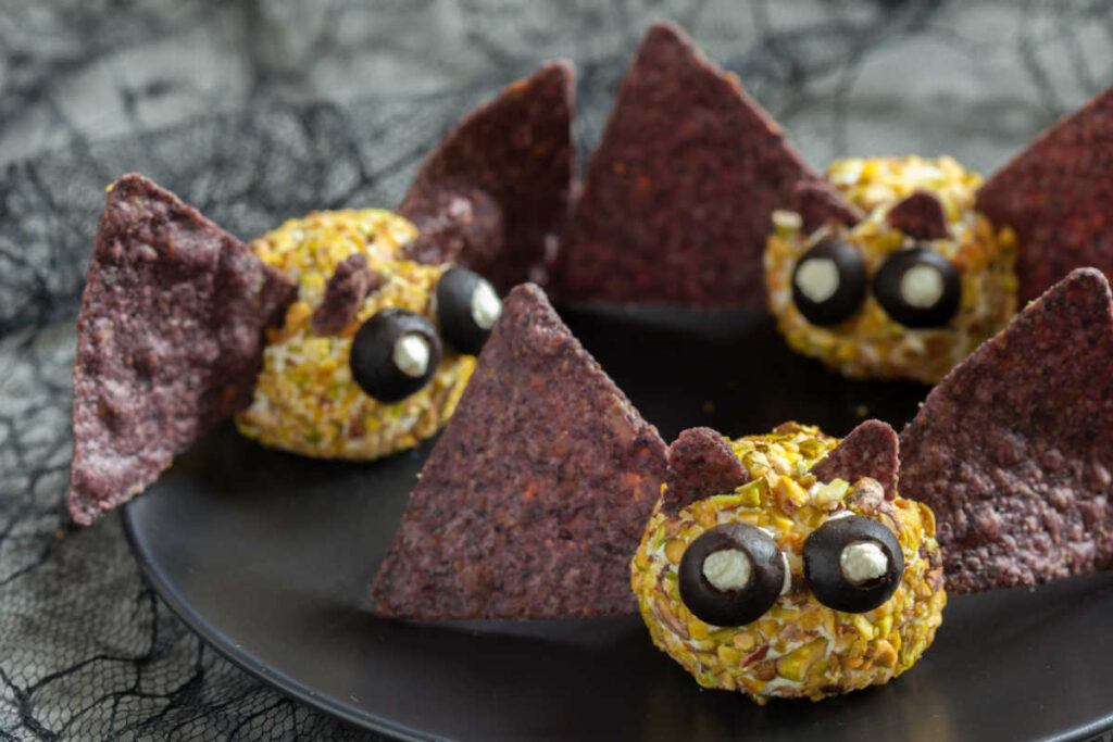 Black plate with 3 Batty Cheese Bites - a creamy cheese ball , rolled in chopped mpistachios and decorated to look like a bat with blue tortilla chi wings and ears. Black olives and goat cheese make the bat's eyes. Side view.