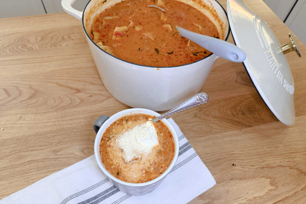 A serving of Lasagna Soup in a gray mug and garnished with a dollop of ricotta cheese and parmesan cheese. Behind the mug is a white pot of lasagna soup with a gray ladle.