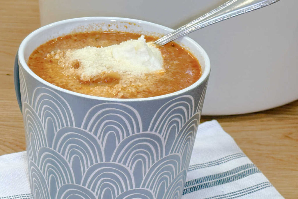 A serving of Lasagna Soup in a gray mug and garnished with a dollop of ricotta cheese and parmesan cheese.