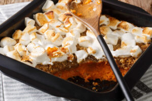 Black casserole dish with a pecan crumble sweet potato casserole. A layer of mashed, spiced sweet potatoes, then a layer of cruncy, sweet, pecan crumble, and topped with marshmallows that are ligtly browned on the top. There is a portion removed with a spoon to show the layers.