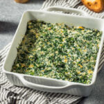White baking dish with creamy, cheesy, Spicy Spinach Madeline, sitting on a gray striped towel.