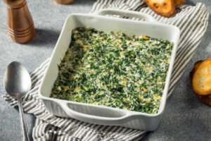 White baking dish with creamy, cheesy, Spicy Spinach Madeline, sitting on a gray striped towel.