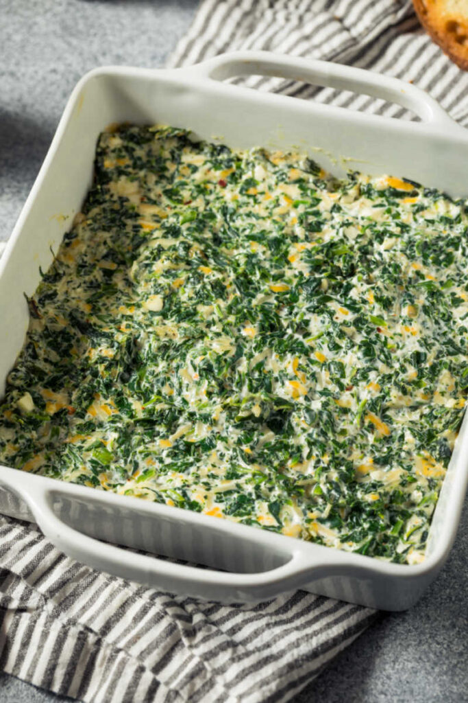 White baking dish with creamy, cheesy, Spicy Spinach Madeline, sitting on a gray striped towel.