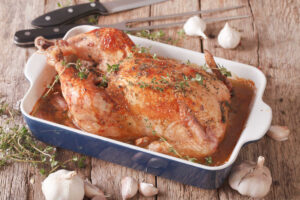 Whole roasted 40 clove garlic chicken in a white and navy blue roaster with garlic cloves and peppercorns laying on a mocha striped tea towel. - topside view.