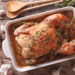 Whole roasted 40 clove garlic chicken in a white and navy blue roaster with garlic cloves and peppercorns laying on a mocha striped tea towel. - overhead view.