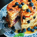 Stack of lemon blueberry goat cheese pancakes with melted butter, fresh blueberries, and a drizzle of syrup, with a wedge removed to see the intrior pancakes. Closeup view.