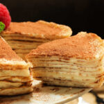 Slice of Stacked Tiramisu Crepe Cake being removed from a whole cake on a wite cake stand.. A cake made of layers of crepes and mascarpone cheese. Closeup view.