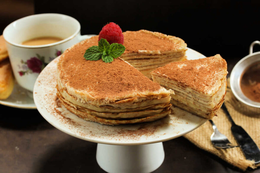 Slice of Stacked Tiramisu Crepe Cake being removed from a whole cake on a wite cake stand.. A cake made of layers of crepes and mascarpone cheese. Side view.