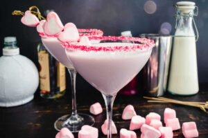 2 Valentine's Day Raspberry White Chocolate Martinis in a martini glass rimmed with red and white sugar and garnished with three small heart marshmallows on a cocktail pick.