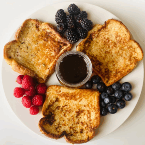 Wite plate with three slices of french toast laying flat on the plate in a circular pattern with a pile of blackberries, a pile of blue berres, and a pile of raspberries beside each slice. There is a small bowl of maple syrup in the center.