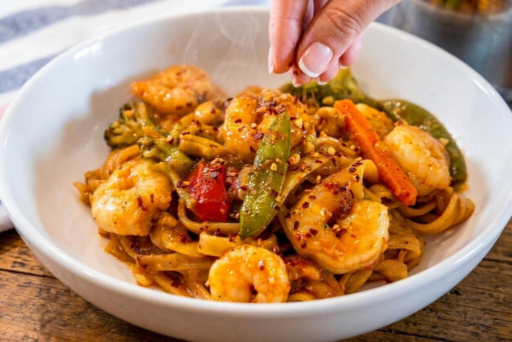 White bowl of steaming Chili Garlic Shrimp And Vegetable Noodles, whith a woman's hand sprinkling red pepper flakes over the top of the food.